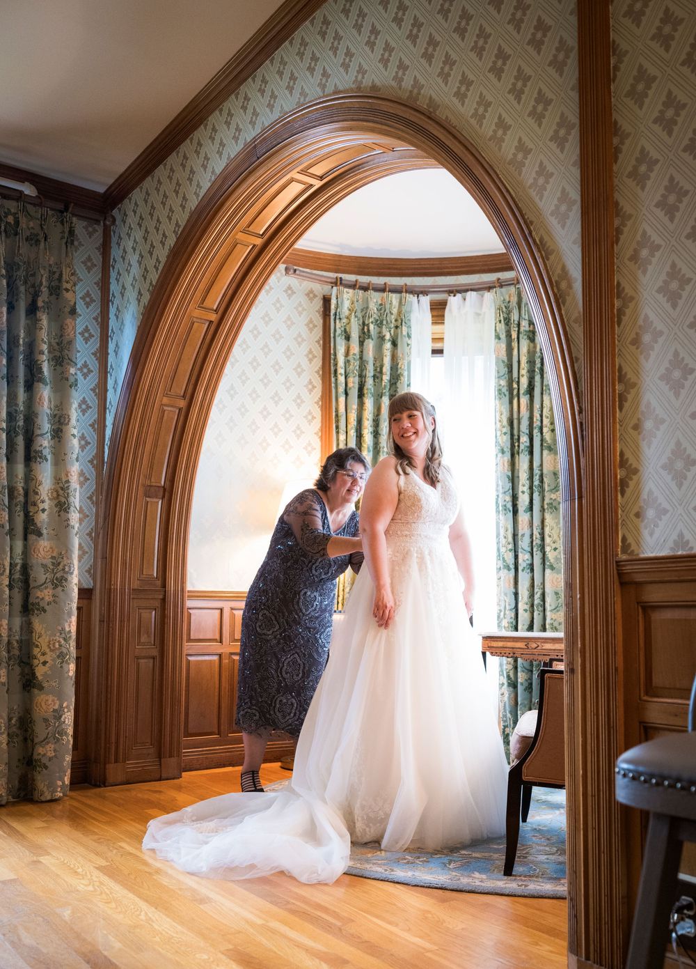 Bride in white gown being helped by woman in blue dress under elegant wood arch in decorated room.