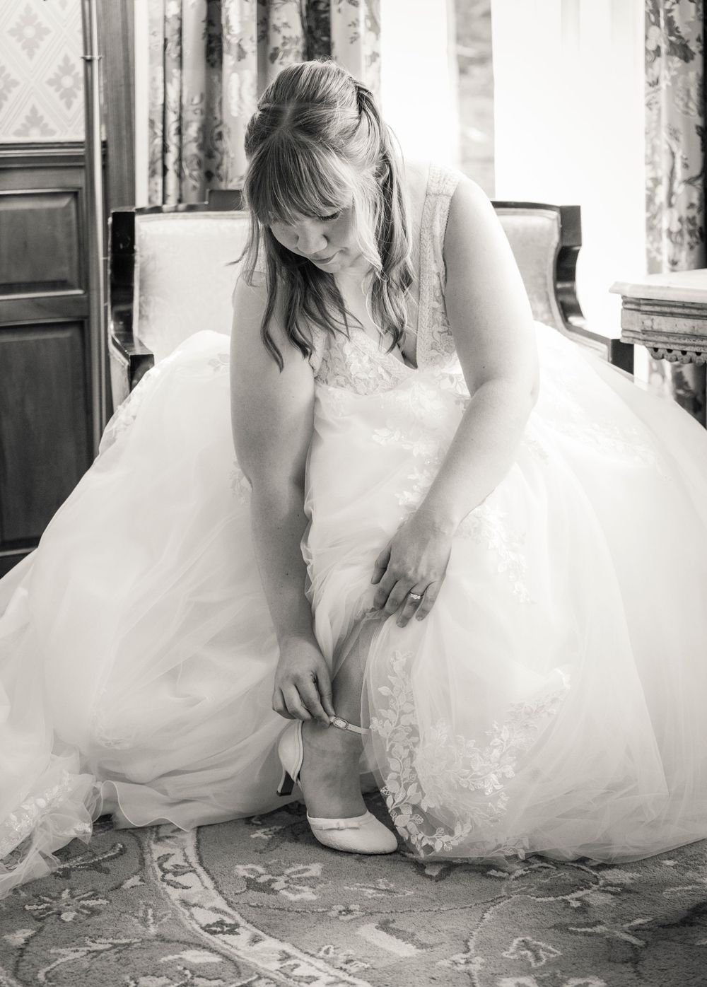 Bride in white wedding gown sitting down putting on her heels in an elegant room, black and white photo.