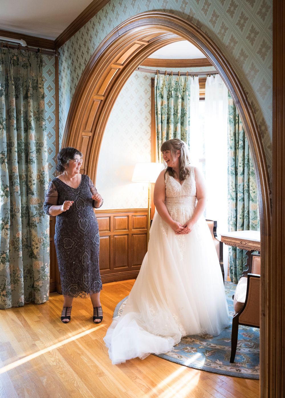 Bride in white gown stands under wooden arch with older woman in blue lace dress in elegant floral-wallpapered room.
