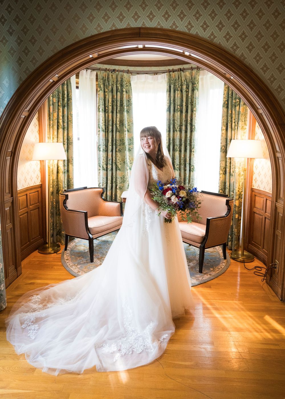 Bride in white gown holds colorful bouquet under elegant wood arch in ornate room with floral curtains.