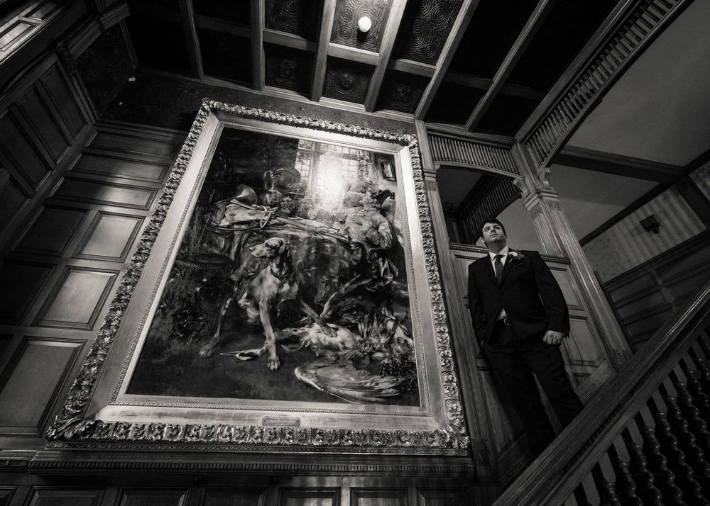 Groom in black suit admiring large ornate painting on staircase wall in black and white photo.