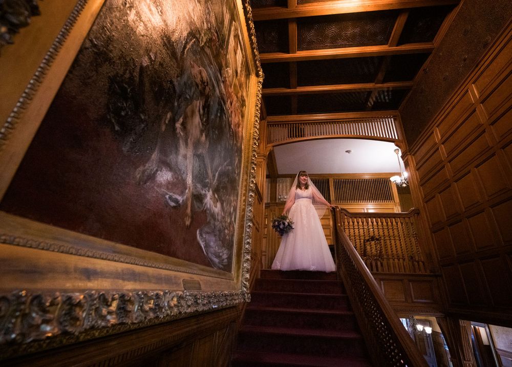 Bride in white gown descends ornate wooden staircase adorned with large classical painting in elegant venue.