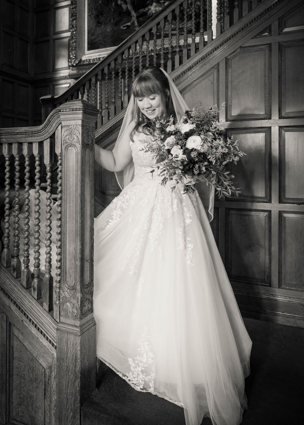 Bride in lace wedding gown holding bouquet on wooden staircase in elegant black and white portrait.