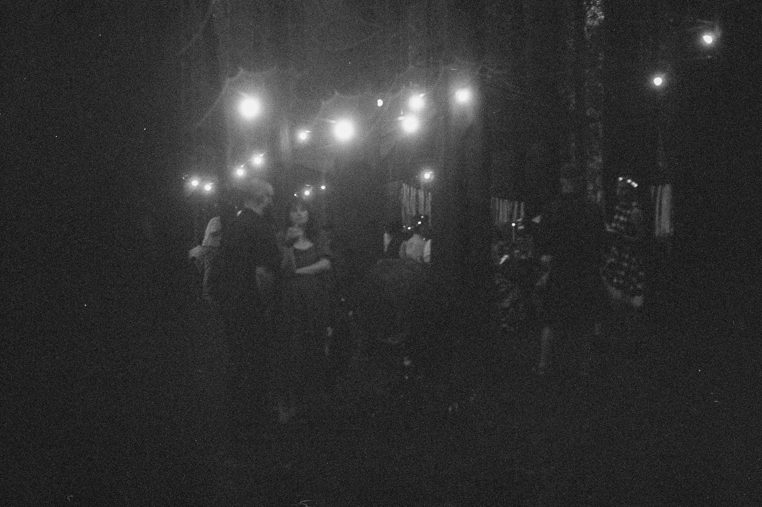 Black and white photo of people gathering outdoors at night under string lights in a dark wooded area.