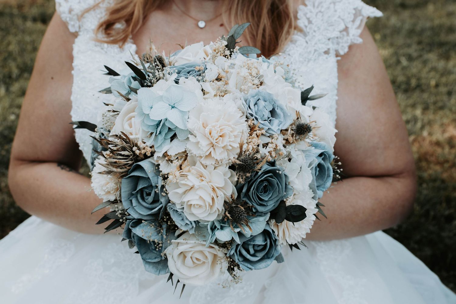 Bride holding a wedding bouquet with dusty blue and cream roses, dried flowers, and greenery accents.
