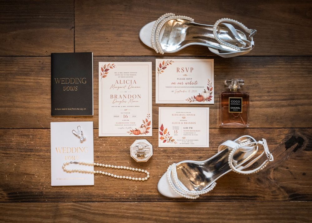 Flat lay of wedding invitation suite, vow book, pearl necklace, perfume bottle and silver heeled sandals on a rustic wooden table.