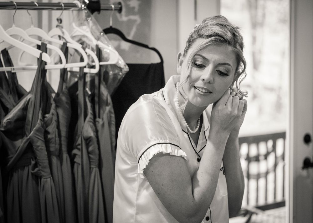Black and white photo of a bride in a satin robe putting on an earring beside bridesmaid dresses hanging on a rack.