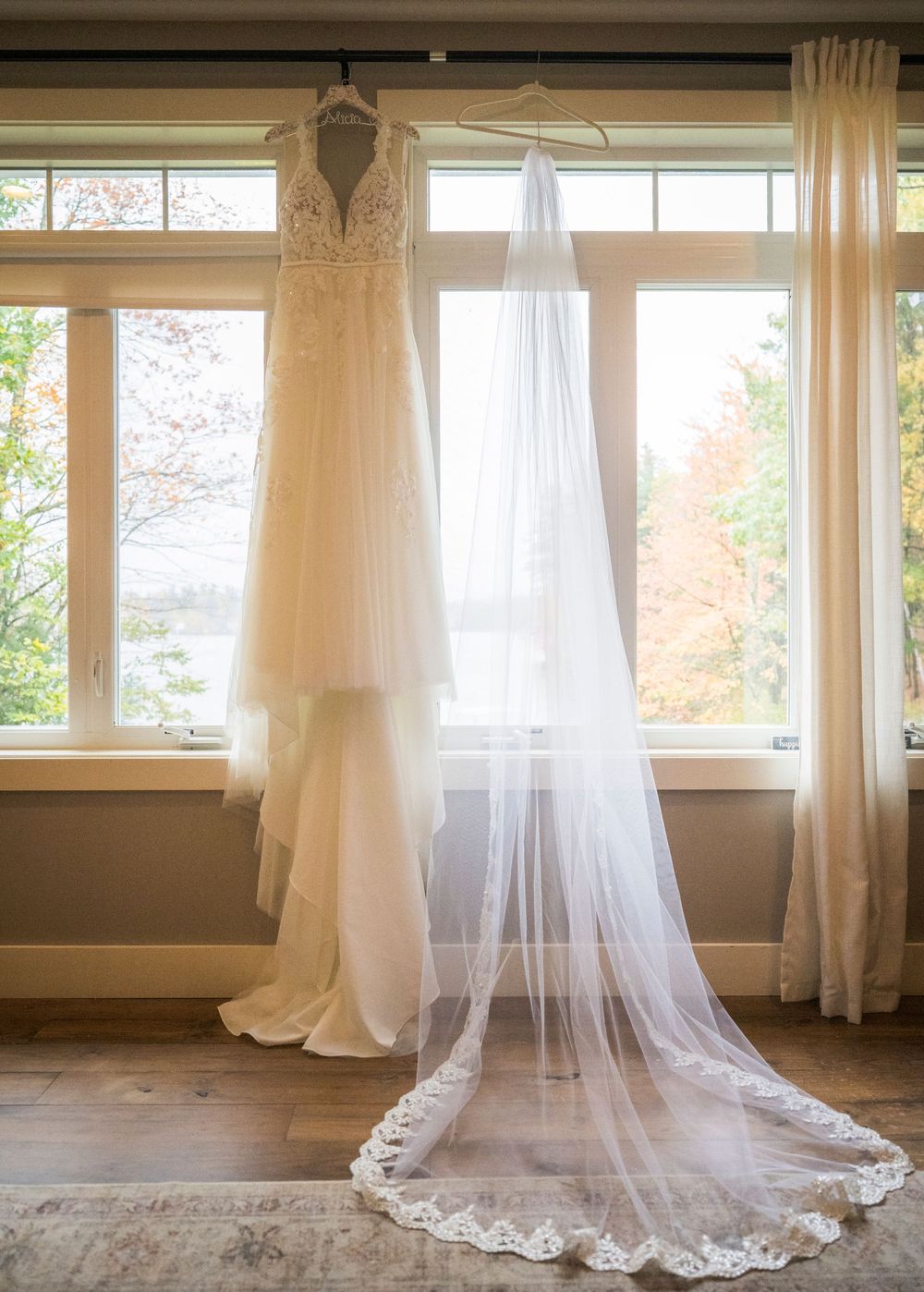 Lace wedding gown and long cathedral veil hanging by large windows overlooking colorful autumn trees.