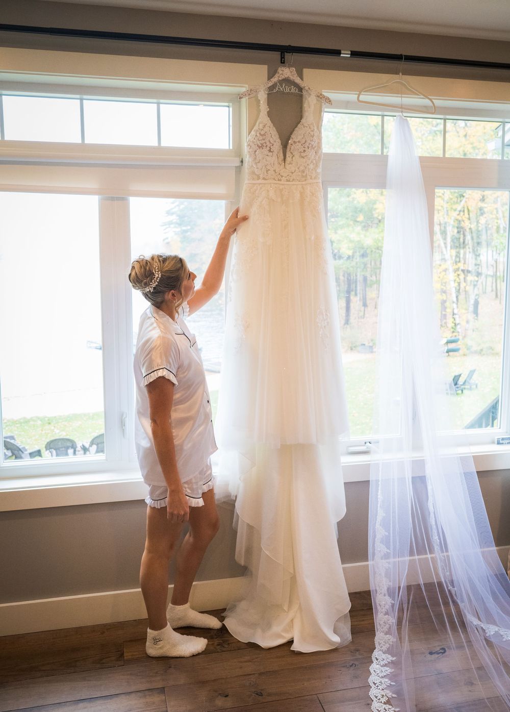 Bride in pajamas reaches up to admire her lace wedding gown and long veil hanging by a bright window before the ceremony.