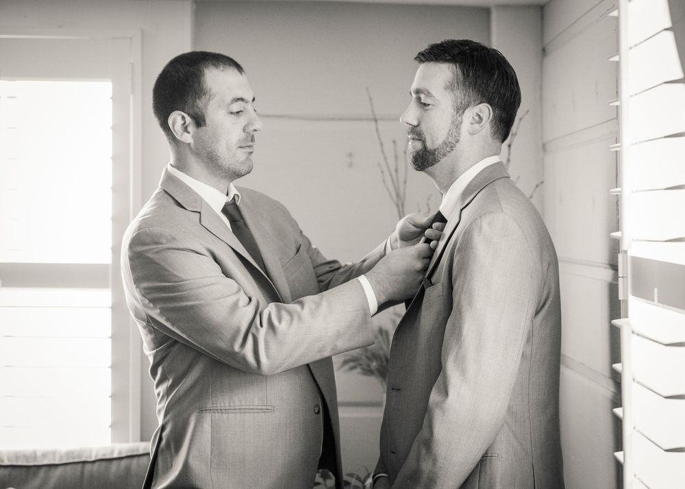 In a black and white photo, a groomsman adjusts the groom's tie as he gets ready for the wedding ceremony in a bright room.