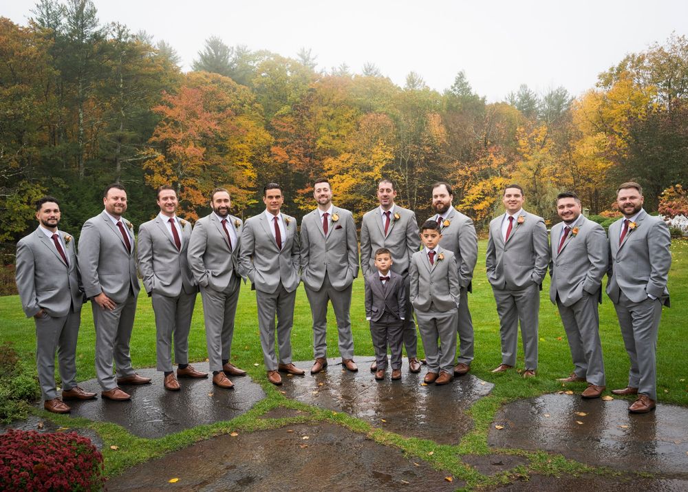 Groomsmen in matching gray suits and burgundy ties pose in a row with a young boy on wet grass, with colorful autumn trees behind.