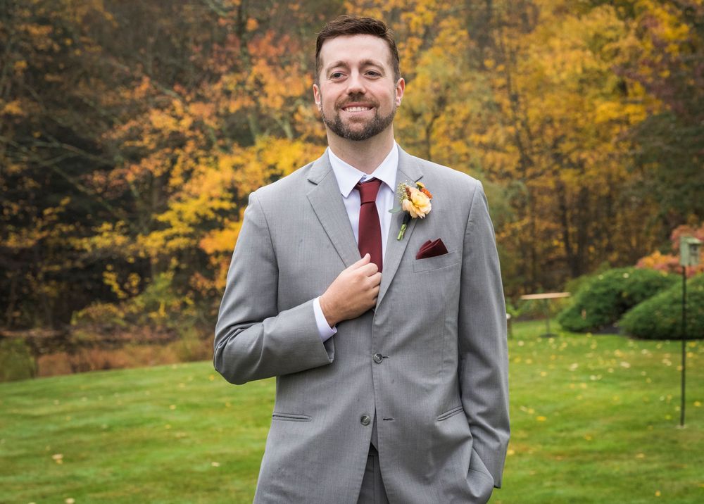 Smiling man in a gray suit with a burgundy tie and yellow boutonniere stands on a green lawn in front of colorful autumn trees.