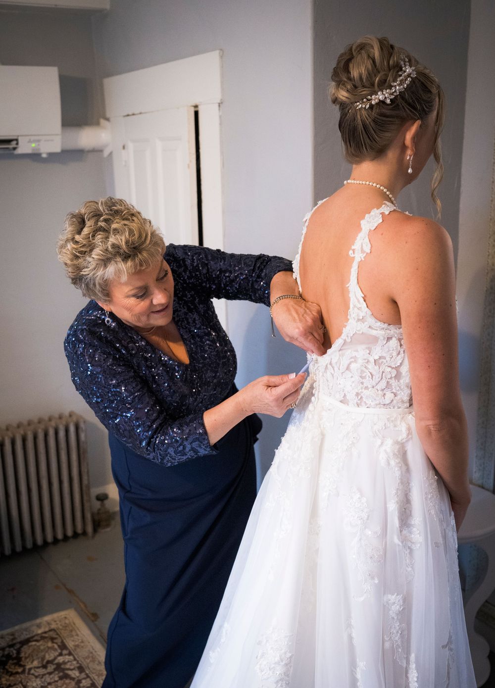 A woman in a navy sequined dress fastens the back of the bride's lace wedding gown while the bride wears a sparkling hair accessory.
