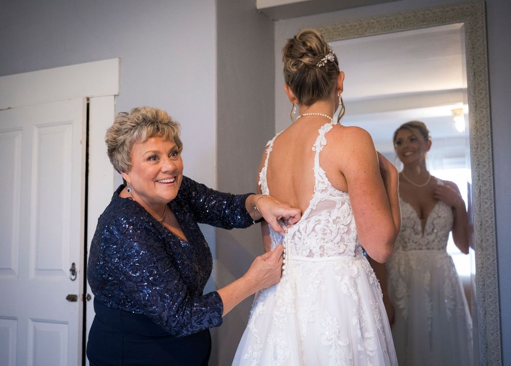 Smiling mother helps the bride button her lace wedding dress while a bridesmaid watches in the mirror.