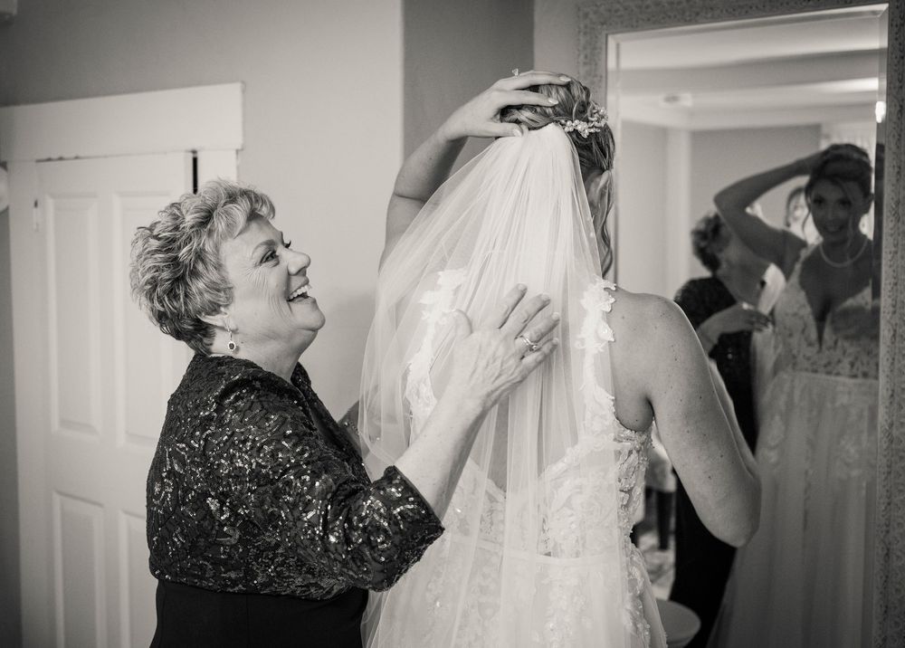 Smiling mother of the bride adjusts the lace wedding veil on the bride as she gets ready in a bright room before the ceremony.