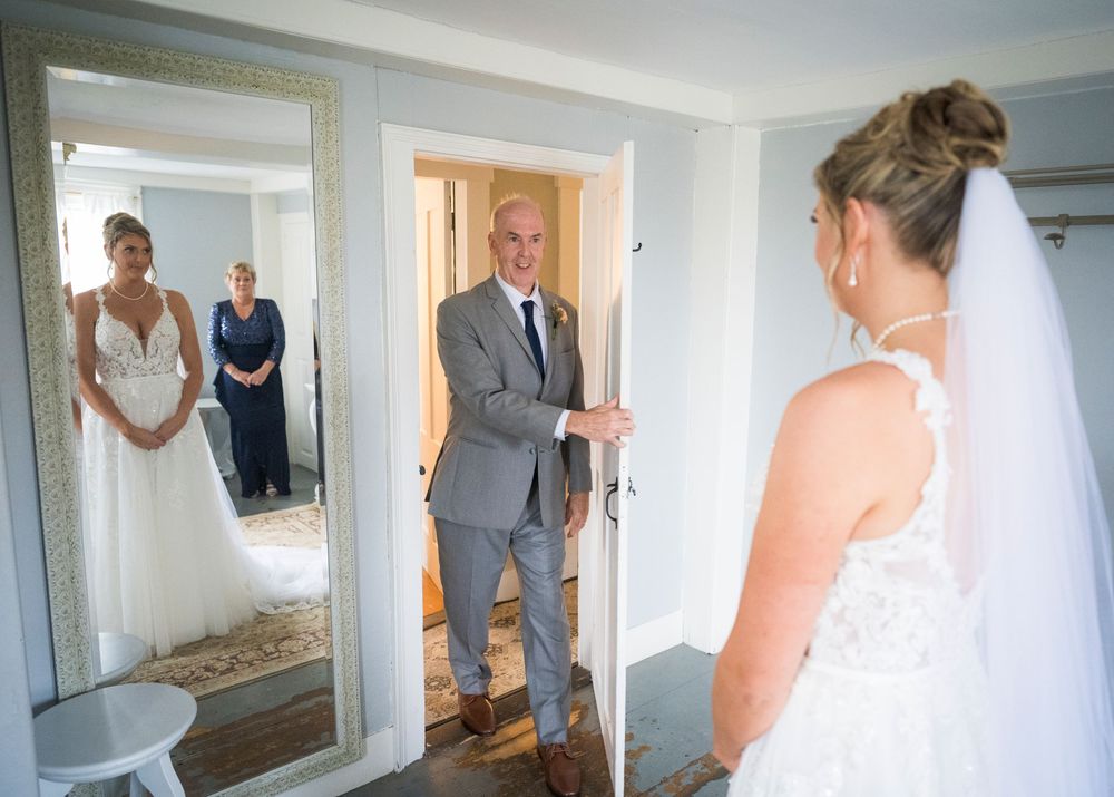A father in a gray suit sees his daughter in her wedding dress for the first time at a doorway, with the moment reflected in a mirror.