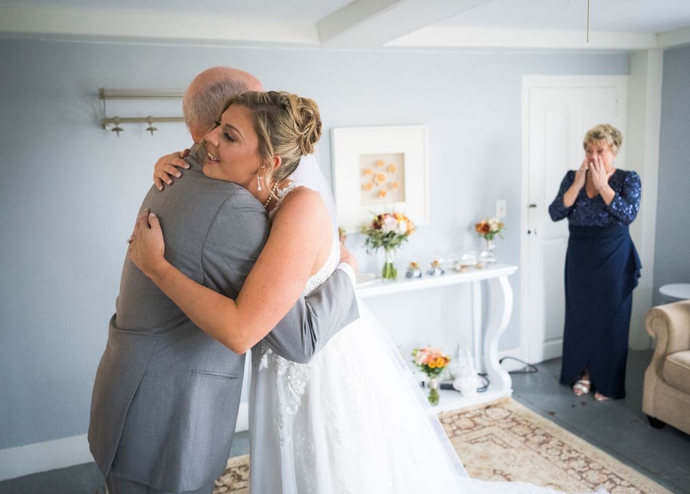 A bride in a white wedding gown hugs an older man in a gray suit while a woman in a navy dress watches emotionally from across the room.