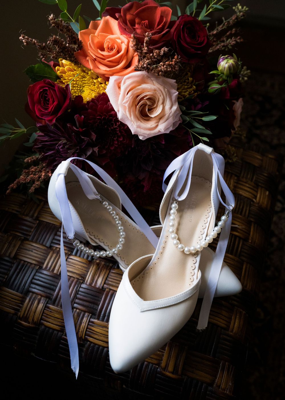 White bridal heels with pearl ankle straps displayed on wicker beside a lush bouquet of red, pink, and orange roses.