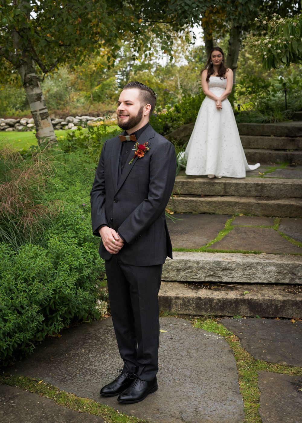 Groom in black suit smiles while bride in white wedding gown stands on stone steps behind him in a lush garden setting.