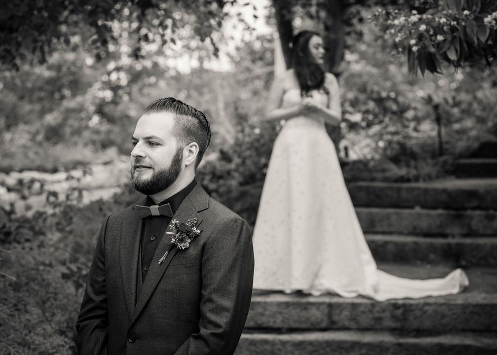 Black and white wedding photo of bearded groom in suit with bride in gown standing on stone steps behind him.