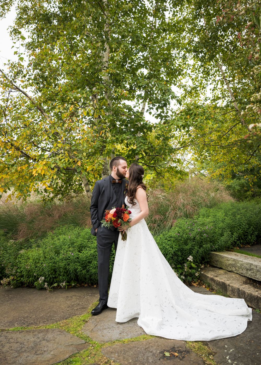 Bride and groom sharing a kiss under a large tree with fall foliage in a garden.