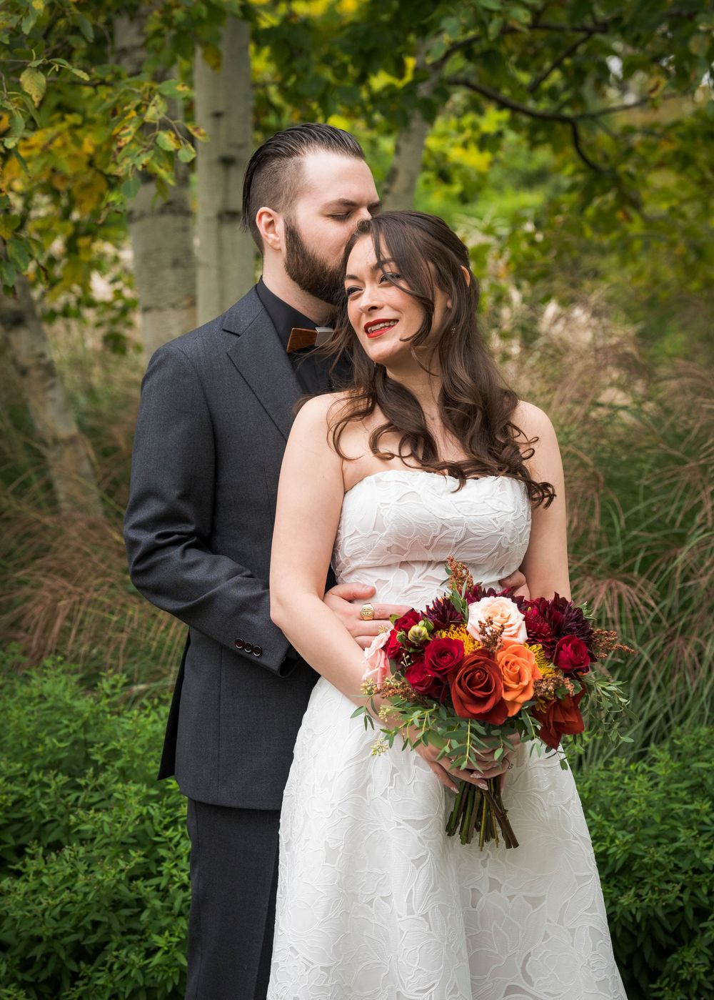 Bride holding autumn bouquet of red and orange roses stands with bearded groom in garden wedding portrait.