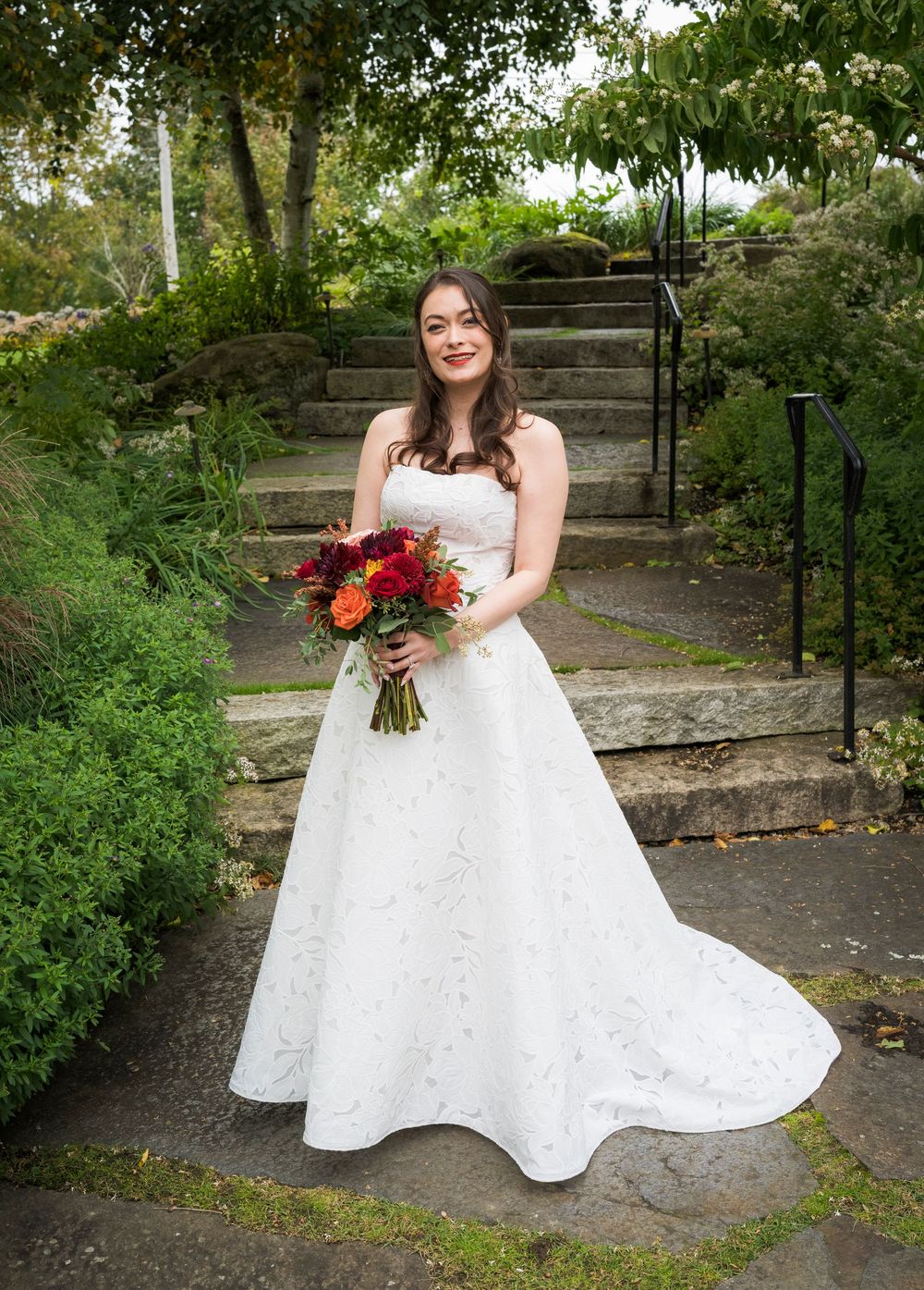 Smiling bride in strapless white gown holding colorful autumn bouquet on garden stone steps.