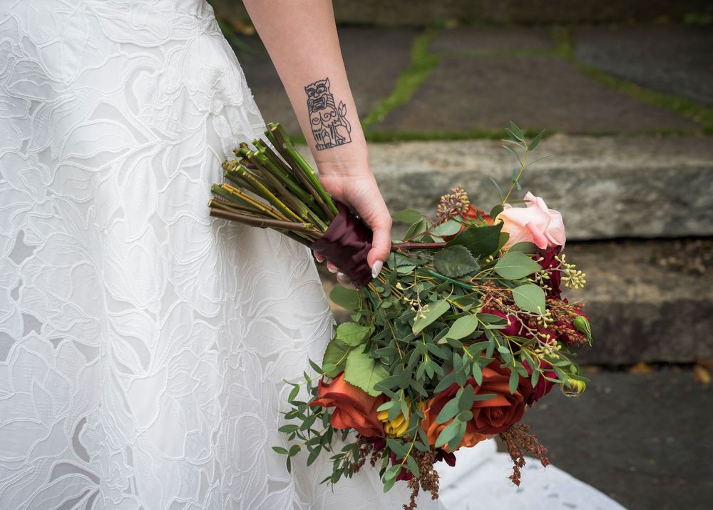 Bride holding a lush fall wedding bouquet of red, orange, and green blooms with a visible tattoo on her wrist.