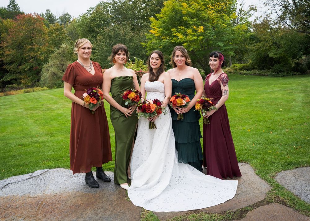 Bride and four bridesmaids in mismatched burgundy and green gowns holding colorful fall bouquets outdoors.