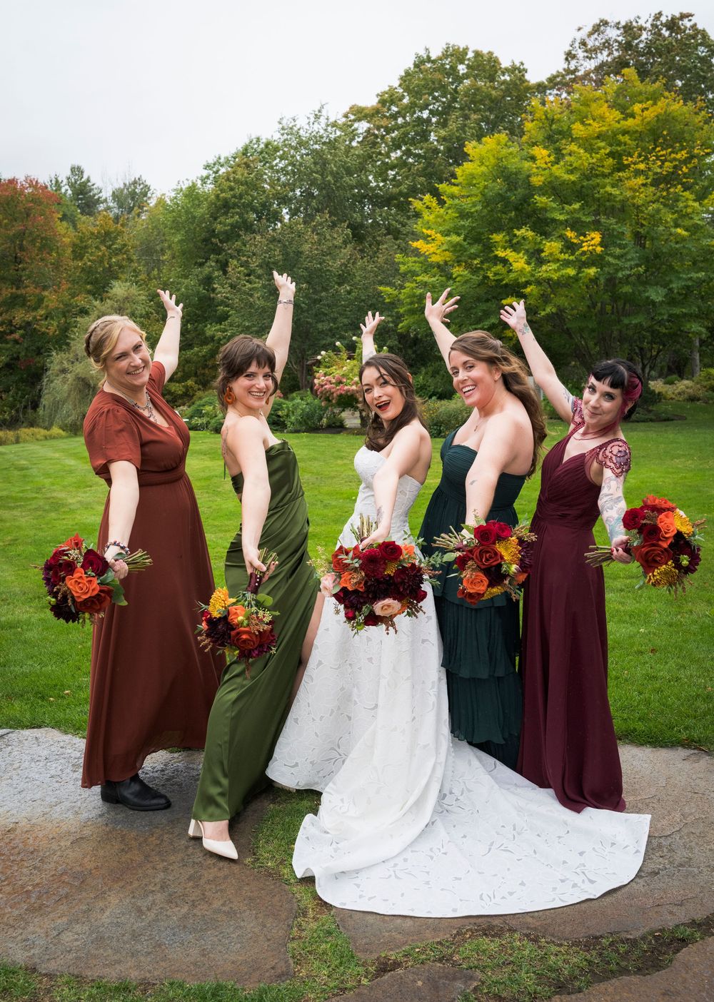 Bride in white lace gown celebrates with four bridesmaids in fall-colored dresses outdoors at autumn wedding.