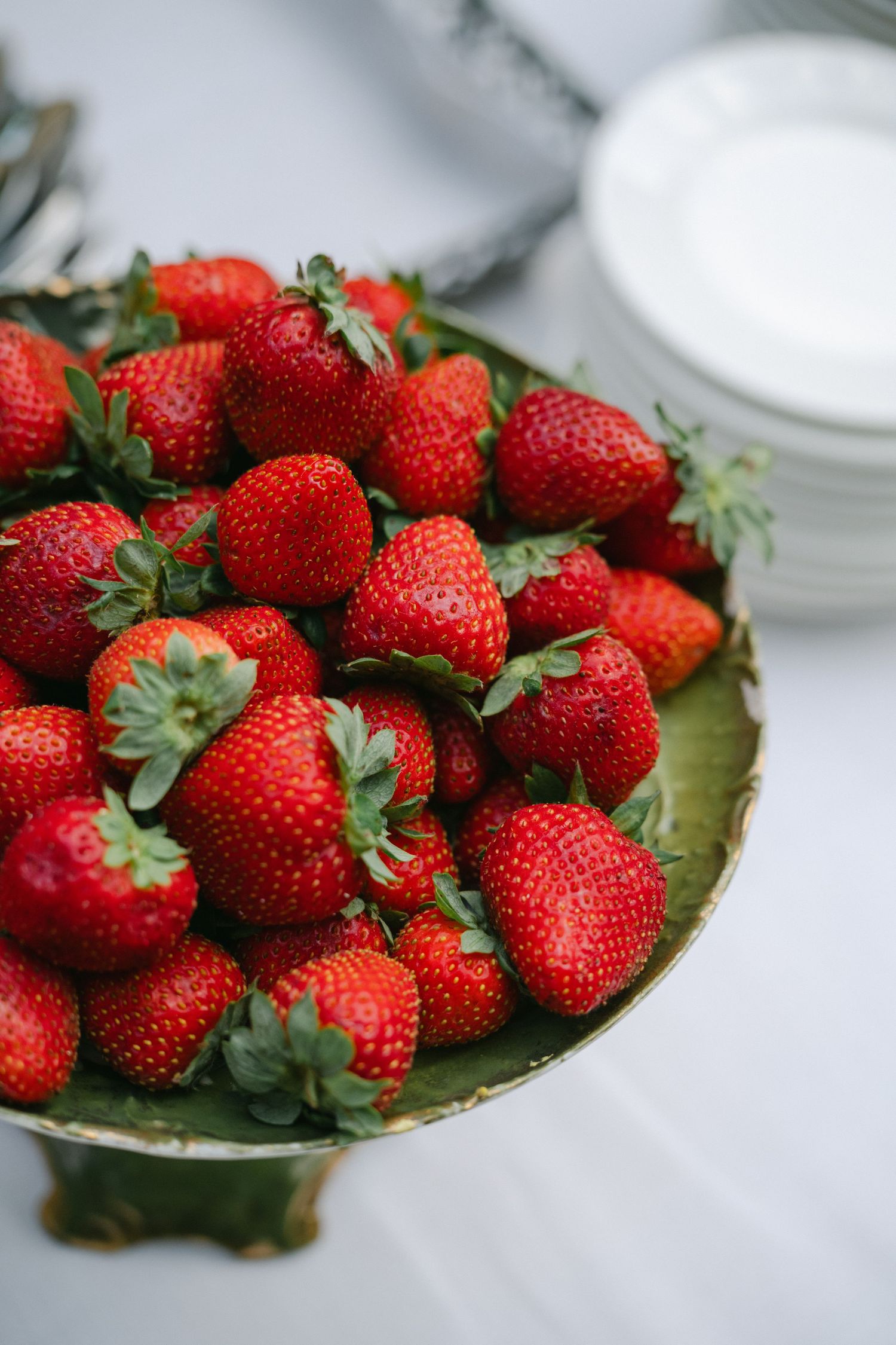 Fresh red strawberries piled high on a vintage green pedestal cake stand, with white plates in the background.