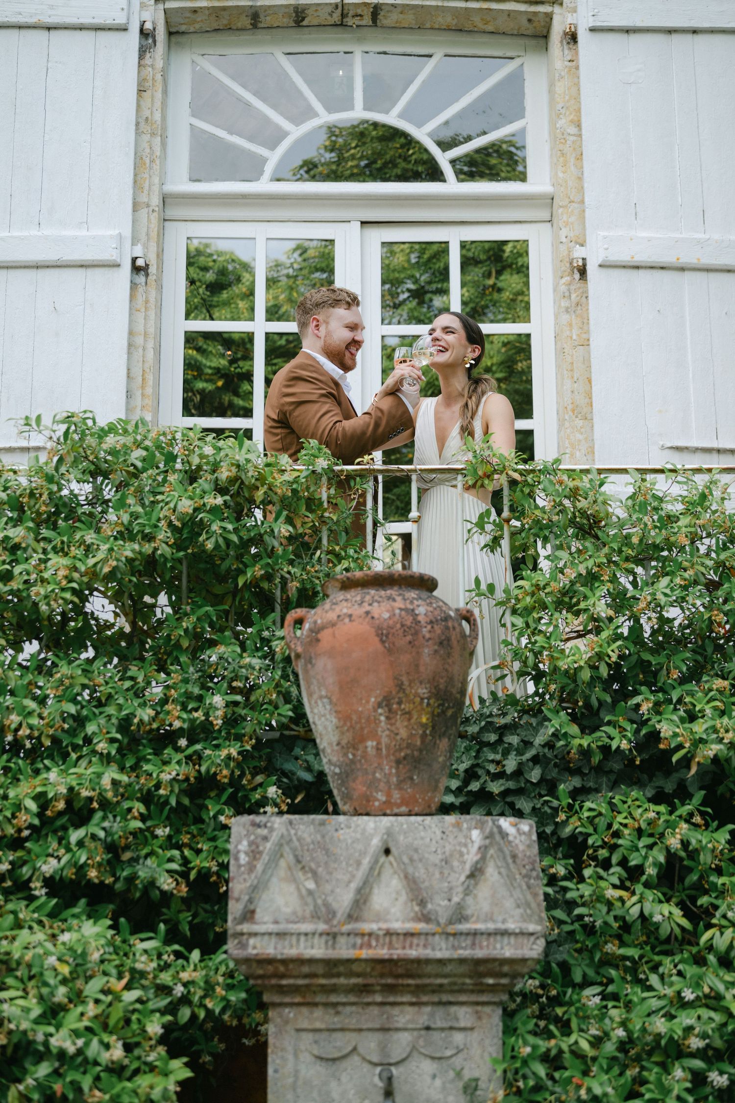 A couple embraces and smiles in front of white gates, groom in brown suit, bride in white pleated dress.