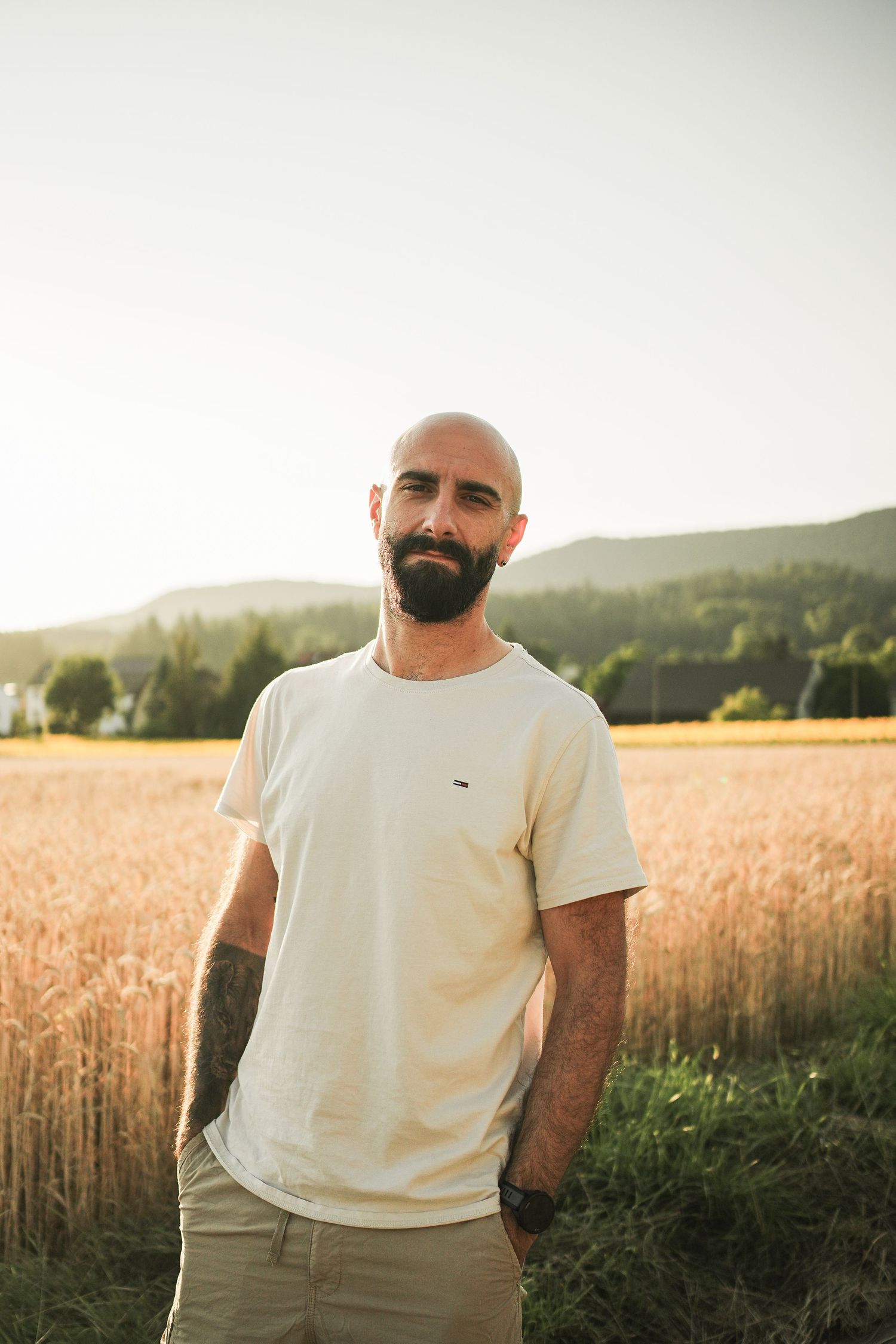 Bald bearded man in white t-shirt standing in golden wheat field at sunset with mountains in background.