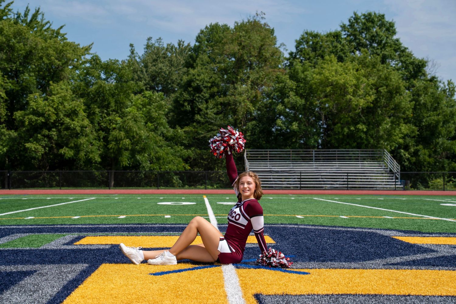 Cheerleader in maroon uniform poses with pom-poms on blue and gold football field turf with bleachers behind.