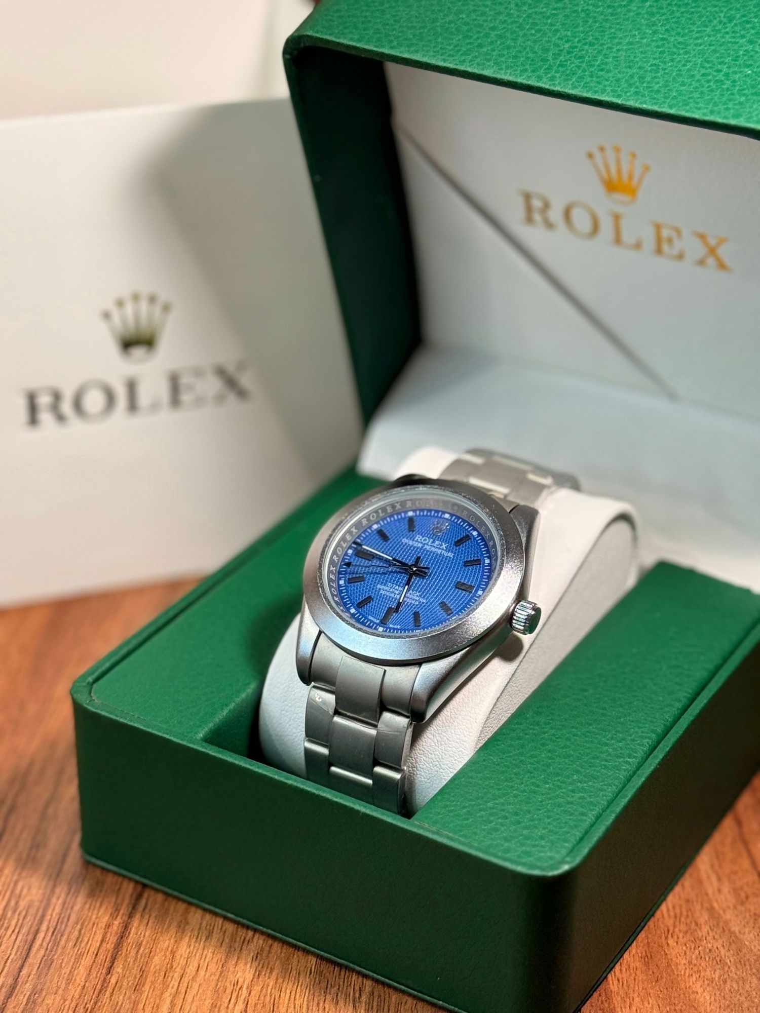Rolex watch with blue dial and stainless steel bracelet displayed in green Rolex box with white cushion.