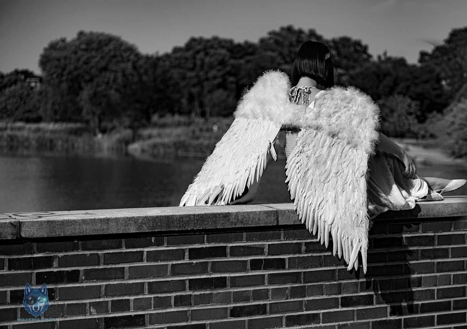 Black and white photo of a person with large white angel wings sitting on a brick wall by a river. Chi.Shooters meet _r.h.folio_