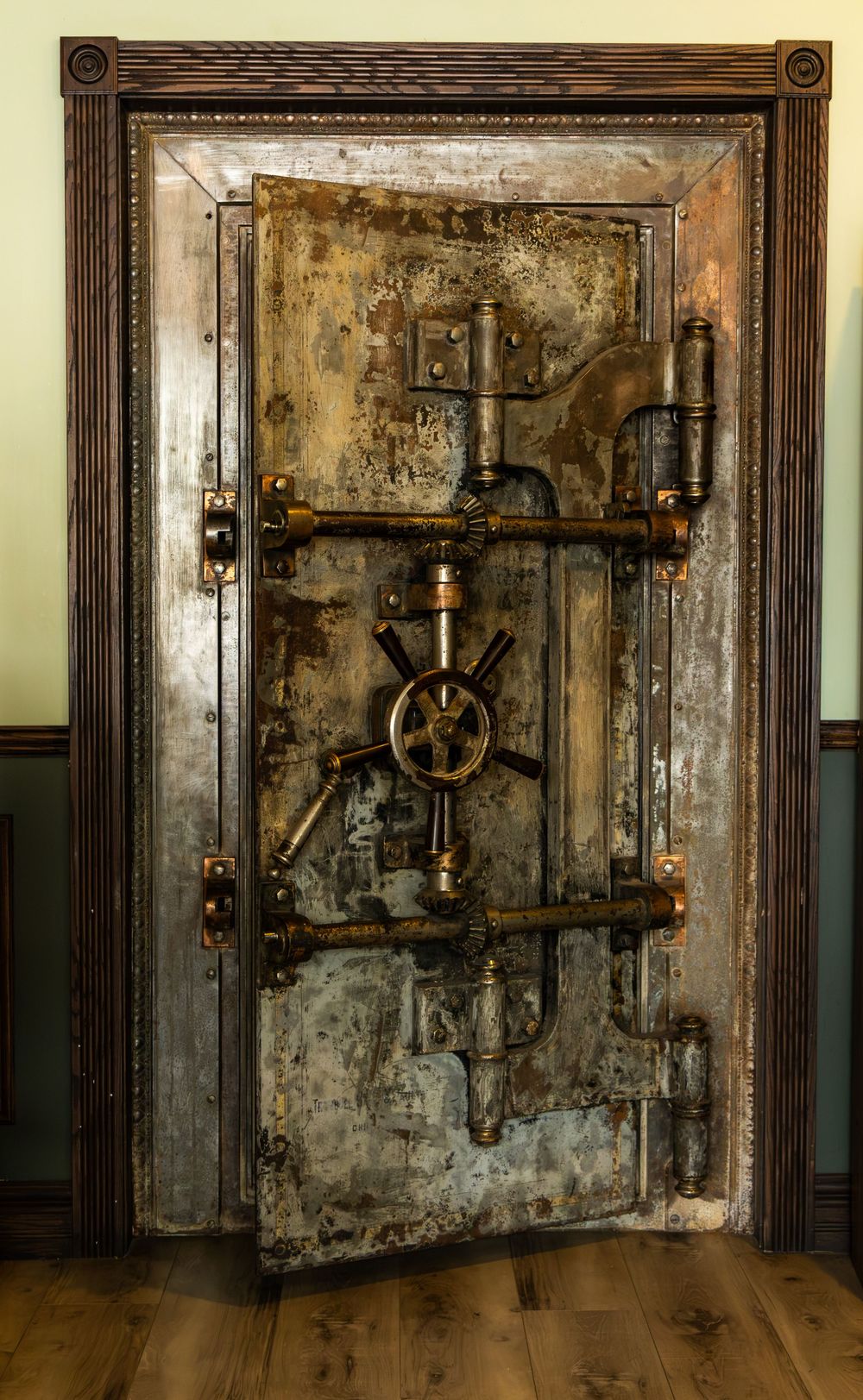 Antique heavy metal bank vault door with brass wheel lock mechanism and ornate wooden frame.