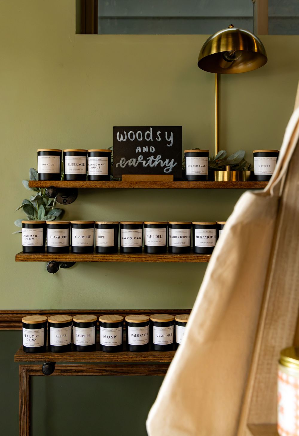 Wooden shelves displaying woodsy and earthy scented candles with gold lids on a sage green wall with a brass lamp.