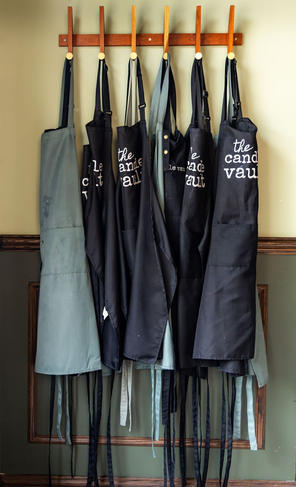 Multiple black and gray aprons labeled 'The Candy Vault' hanging on a wooden wall-mounted coat rack.