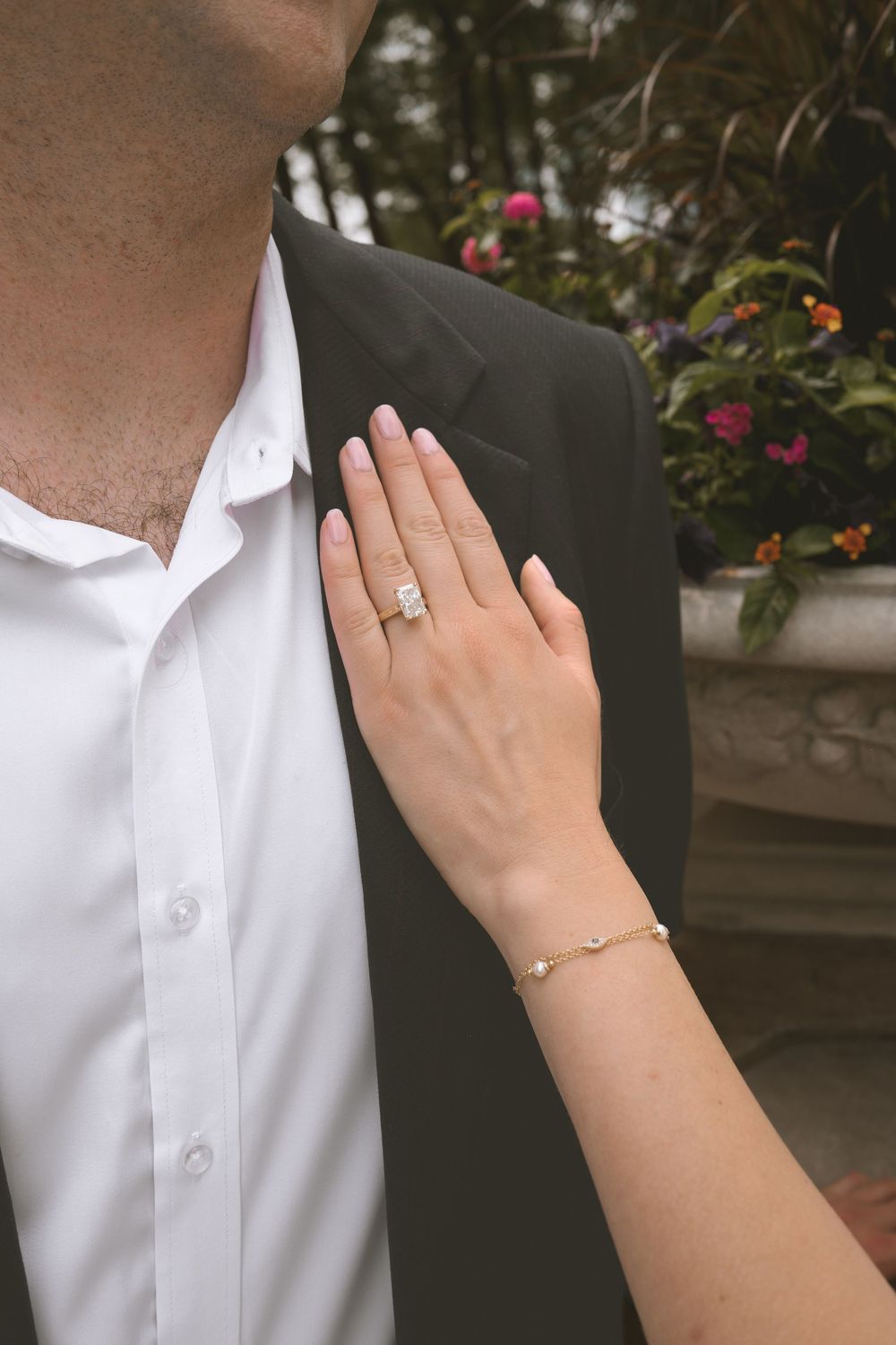Close-up of a woman's hand with diamond engagement ring and gold bracelet resting on a man's dark suit jacket.