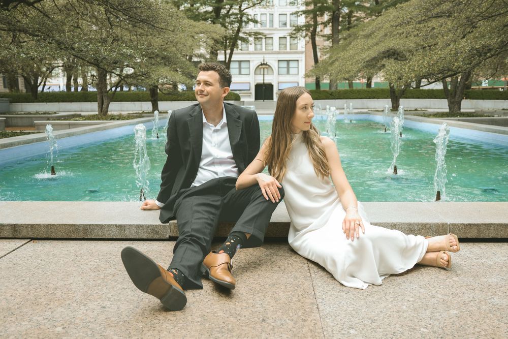 A couple sits casually by an outdoor fountain in a courtyard, man in dark suit, woman in white dress.