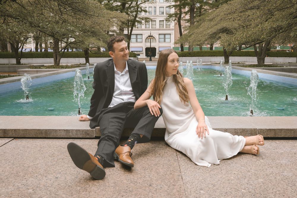 Couple sitting together by an outdoor fountain in a park, man in dark suit and woman in white dress.