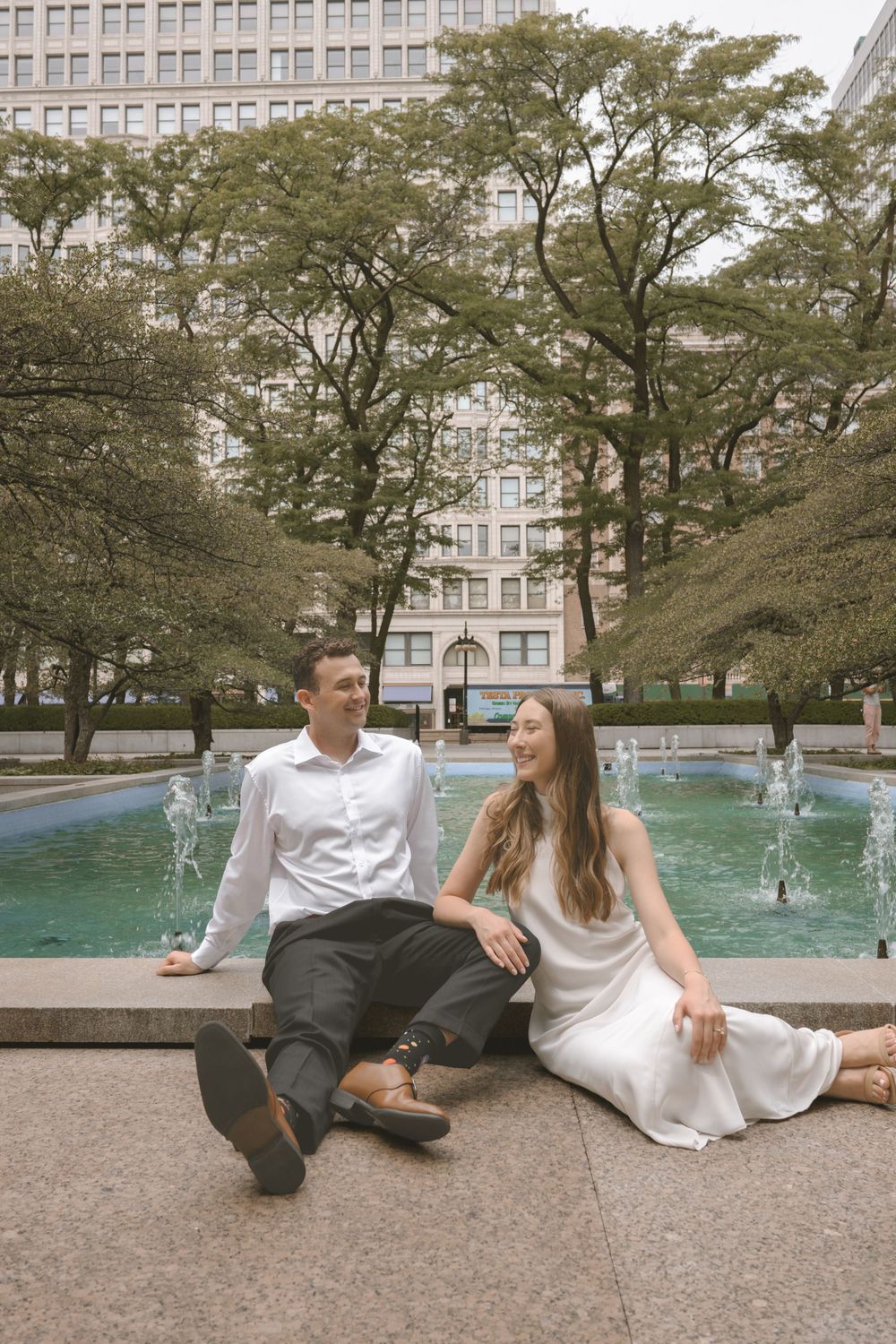 Couple sitting by a city fountain, woman in white dress and man in white shirt, surrounded by trees and urban buildings.