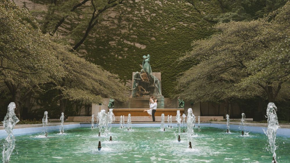 Ornate fountain with multiple water jets in a lush green park featuring a classical statue monument surrounded by trees.