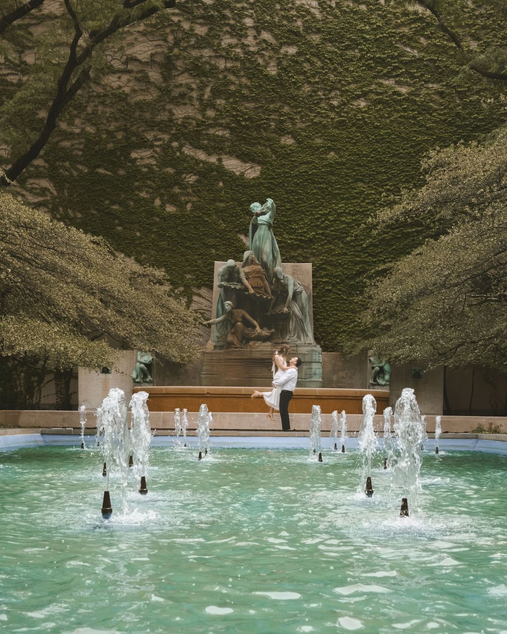 Couple embracing near a fountain with bronze statues and ivy-covered walls in a lush garden setting.