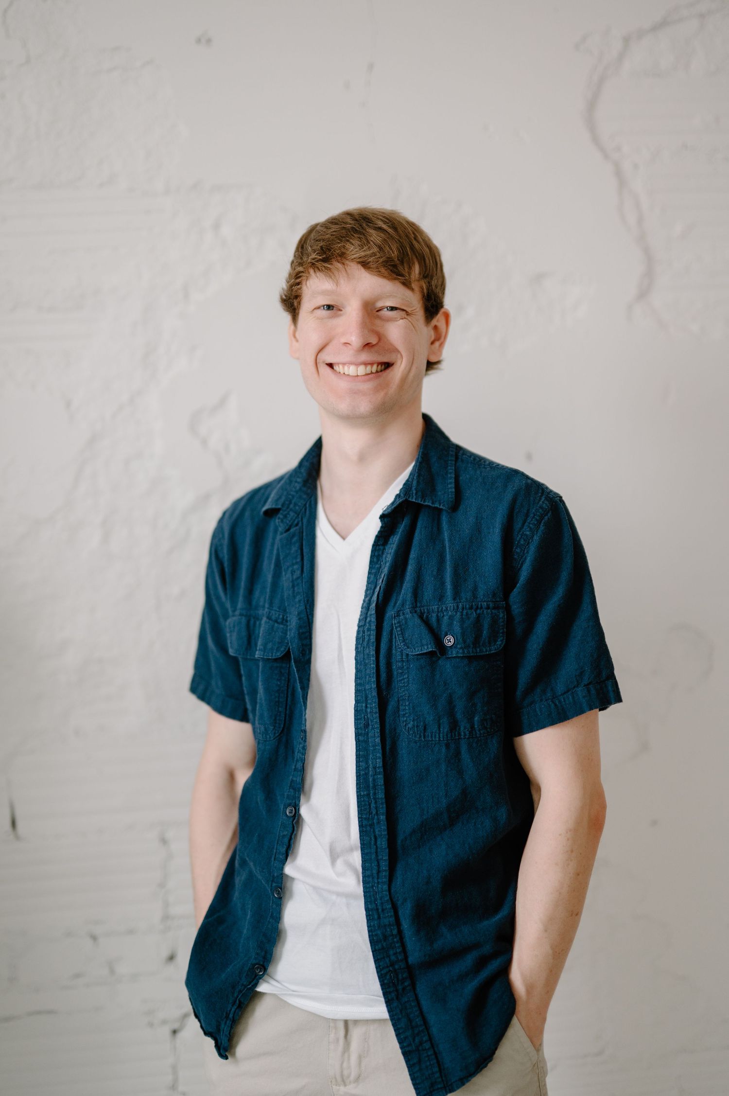Smiling young man with red hair wearing a navy shirt over white tee, standing against a white brick wall.