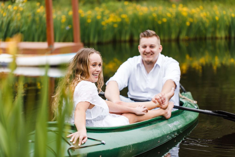 Smiling couple enjoying a romantic canoe ride on a calm lake surrounded by lush green trees and yellow wildflowers.