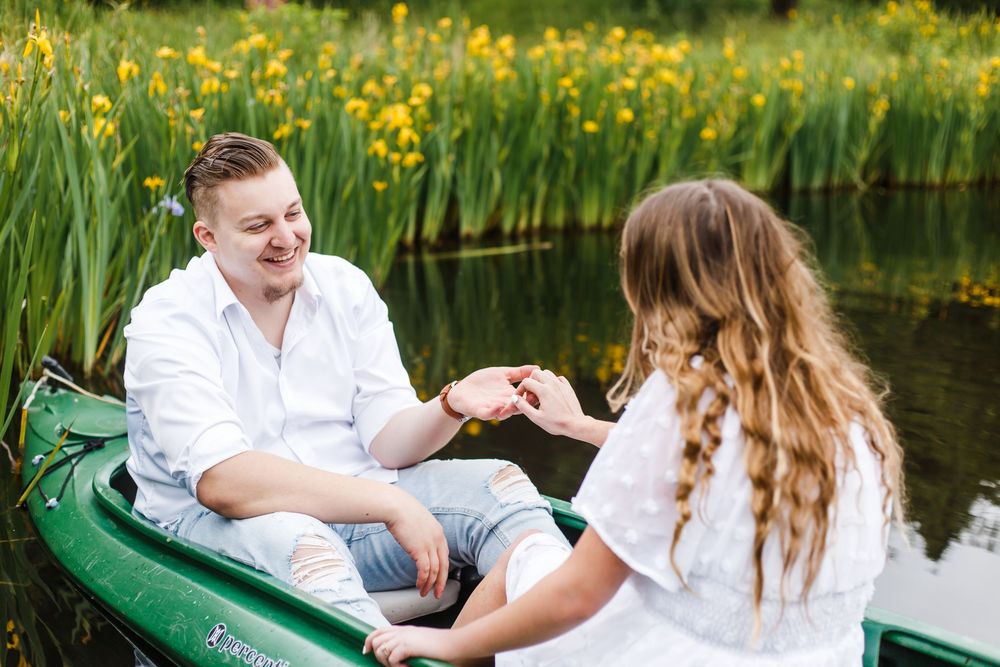 Couple holding hands in a green rowboat on a calm pond surrounded by yellow wildflowers and lush greenery.