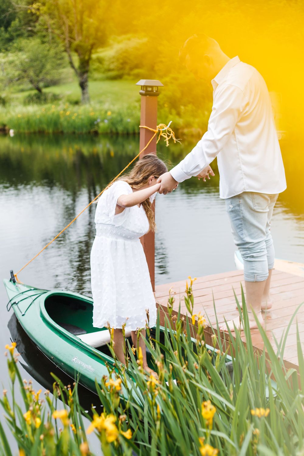 Father and daughter standing on a wooden dock by a calm lake, next to a green canoe, surrounded by yellow iris flowers.