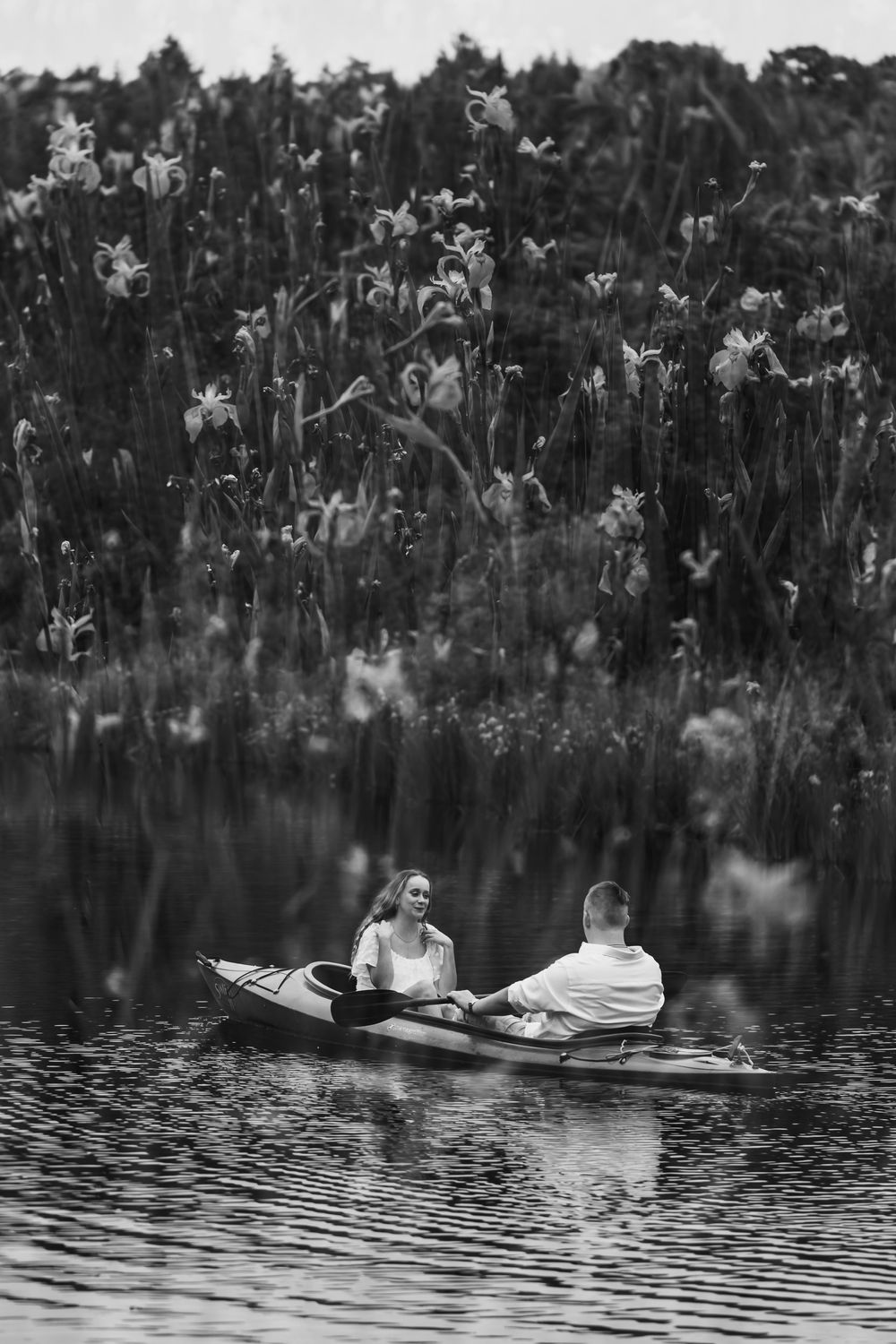 Black and white photo of a couple rowing a kayak on a calm lake surrounded by blooming wildflowers.
